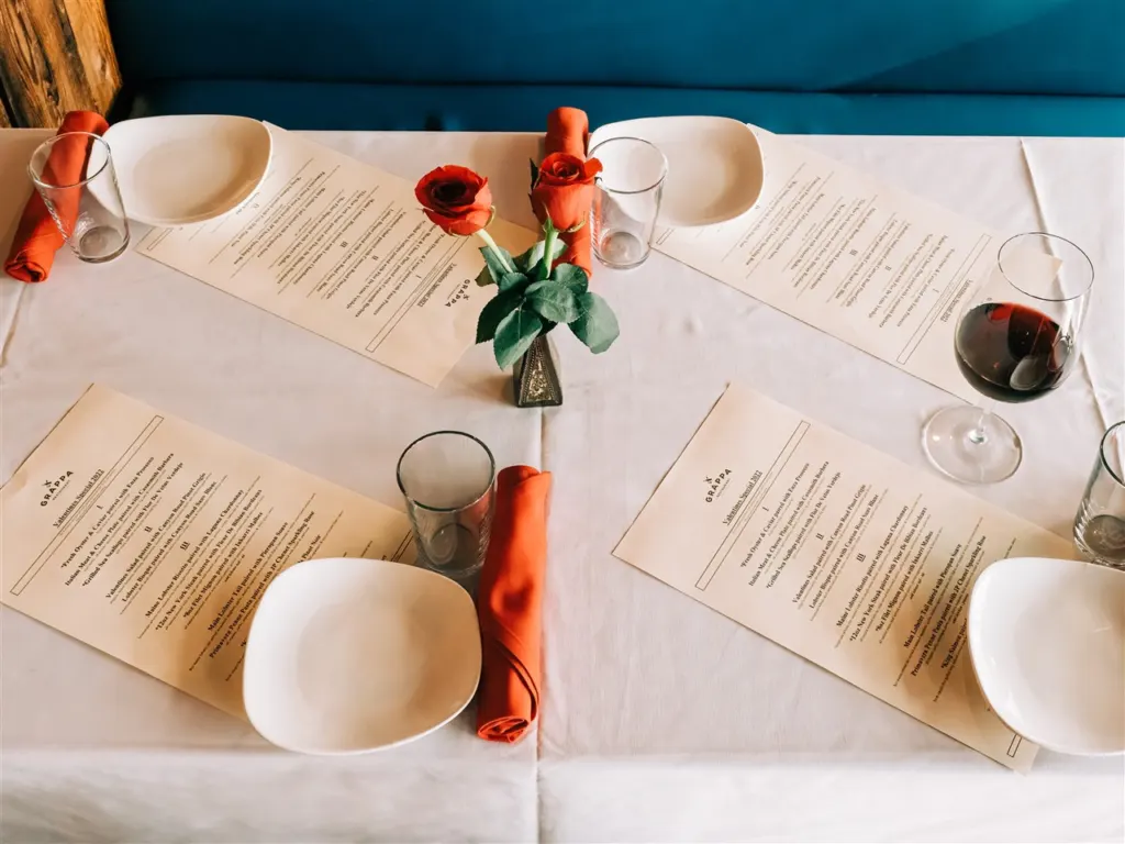 A table at Grappa in Queen Anne set with printed menus, white plates, orange napkins, red roses and a glass of red wine, before Converge at El Corazon