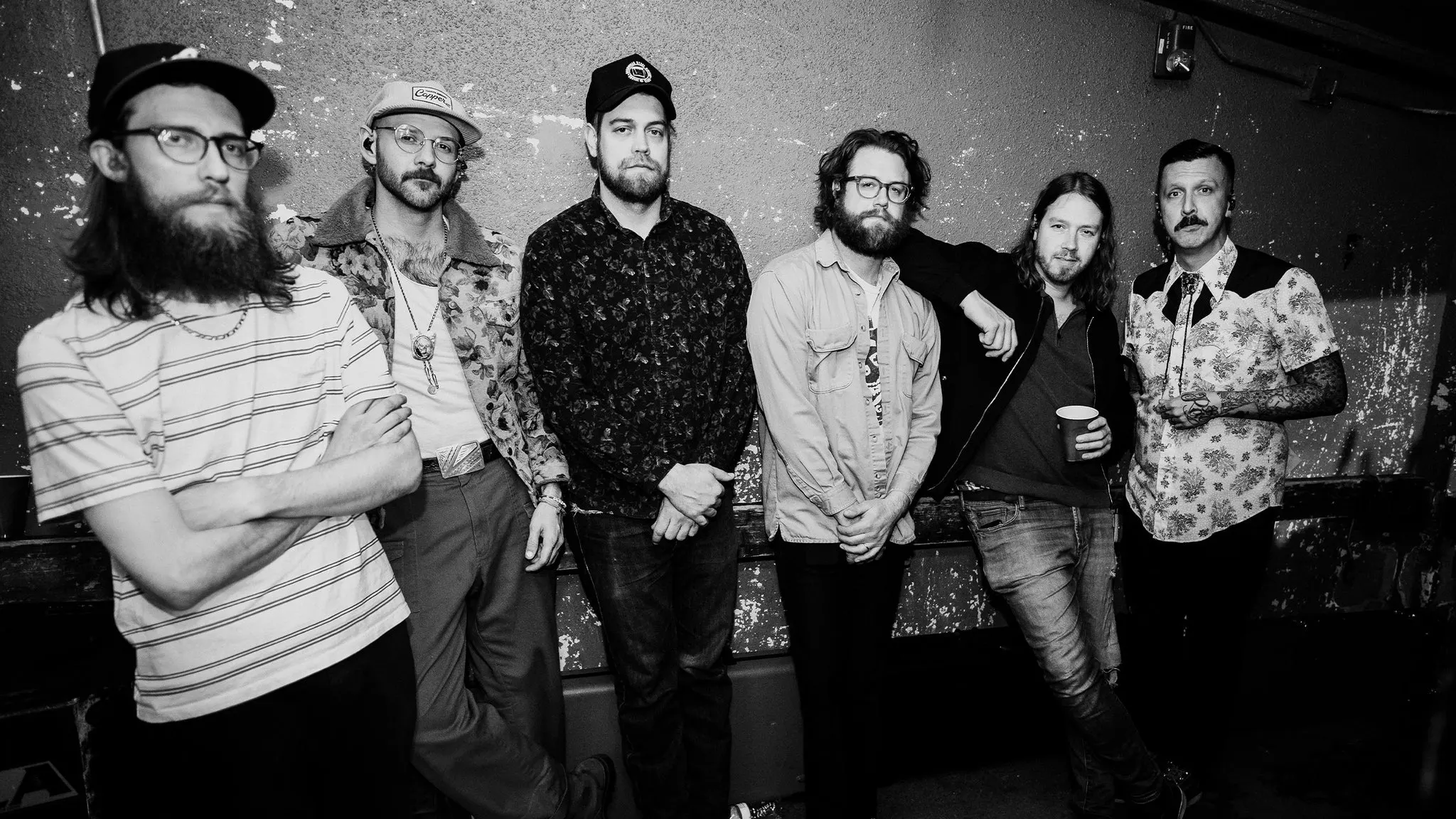Black and white photo of the six members of American Aquarium standing against a weathered, peeling wall, ahead of their show at The Crocodile near Grappa