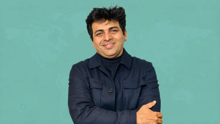 Comedian Amit Tandon smiling with his arms folded in a navy jacket in front of a teal world map, ahead of Halwa at the Moore Theatre near Grappa in Queen Anne