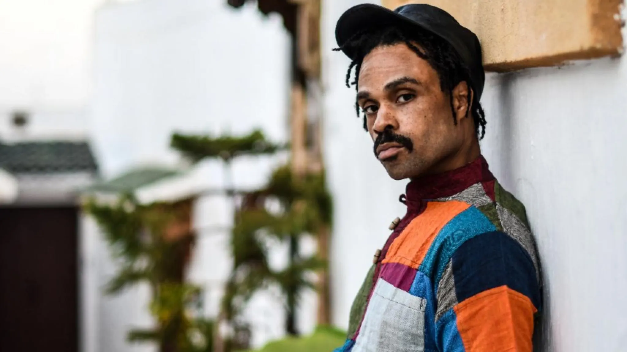 Singer Bilal in a black cap and a colorful patchwork jacket leaning against a white wall, ahead of his show with Yaya Bey at The Crocodile near Grappa