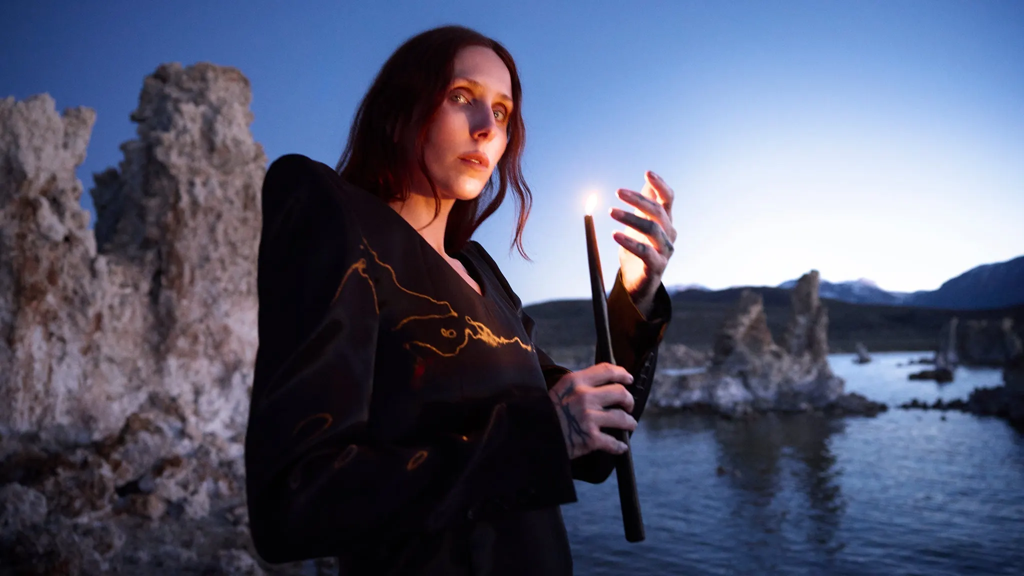 Chelsea Wolfe in an embroidered black jacket holding a lit candle beside rocky tufa towers and a lake at dusk, ahead of her Moore Theatre show near Grappa in Queen Anne