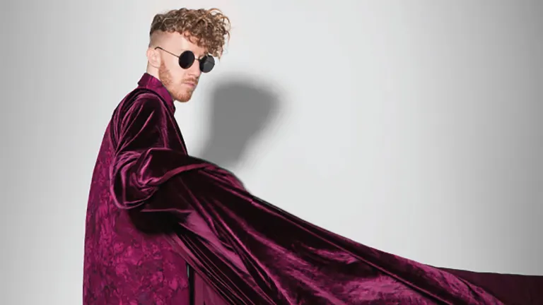 Singer Daley in round dark sunglasses and a flowing burgundy velvet coat against a pale wall, ahead of his Nectar Lounge show near Grappa