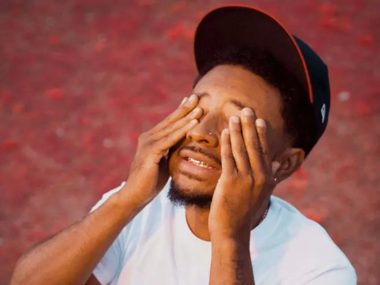 Rapper Deante' Hitchcock in a white T-shirt and black cap covering his eyes with both hands and smiling, ahead of his Hidden Hall show near Grappa