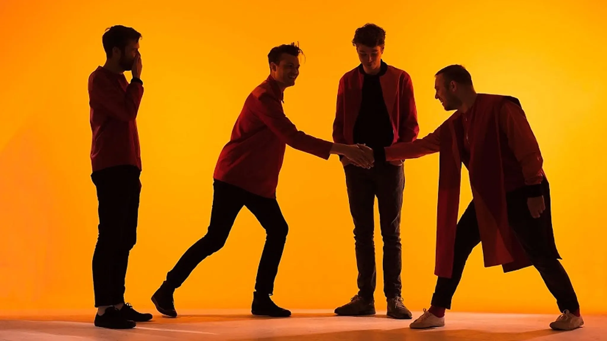 Silhouettes of the four members of Everything Everything in dark red clothes against a bright orange backdrop, two shaking hands, ahead of their show at The Crocodile near Grappa