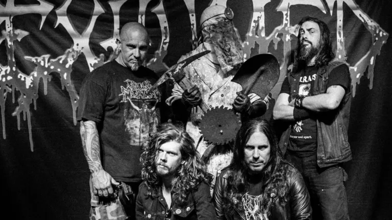 Black and white photo of Exhumed posing in front of a dripping band banner beside a masked figure holding a pickaxe, a shovel and a saw blade, ahead of their El Corazon show near Grappa