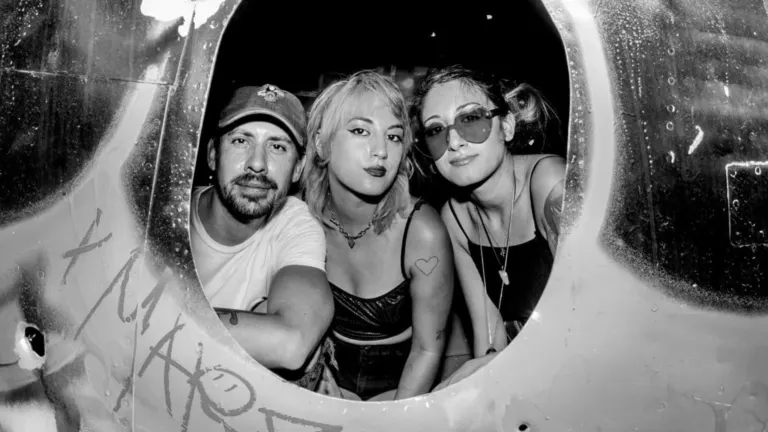 Black and white photo of three members of Eyes Set To Kill peering out through an oval opening in a graffiti-marked metal wall, ahead of their El Corazon show near Grappa
