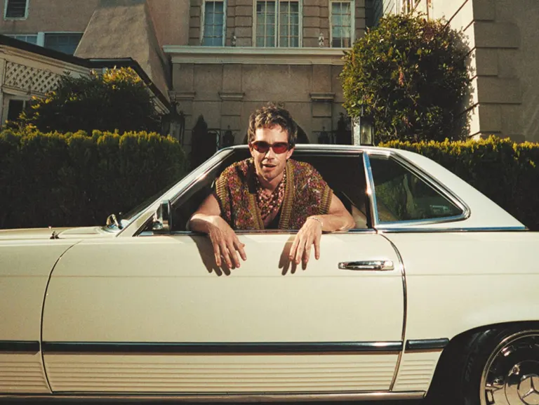 Geordie Kieffer in red sunglasses and a patterned knit shirt leaning out of the window of a vintage cream convertible, ahead of his Hidden Hall show near Grappa