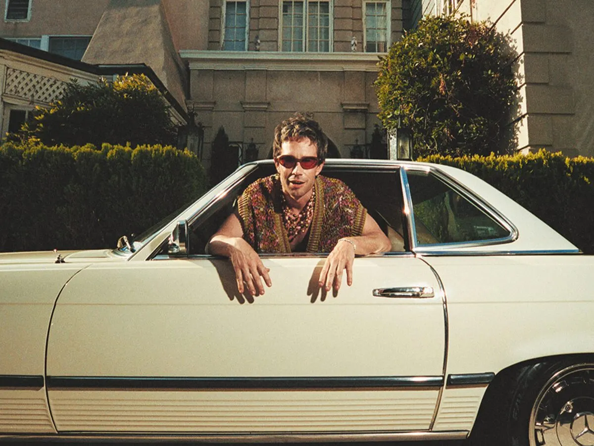 Geordie Kieffer in red sunglasses and a patterned knit shirt leaning out of the window of a vintage cream convertible, ahead of his Hidden Hall show near Grappa