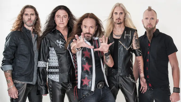 The five members of HammerFall in studded leather and denim posing against a white wall, the singer reaching toward the camera, ahead of their El Corazon show near Grappa