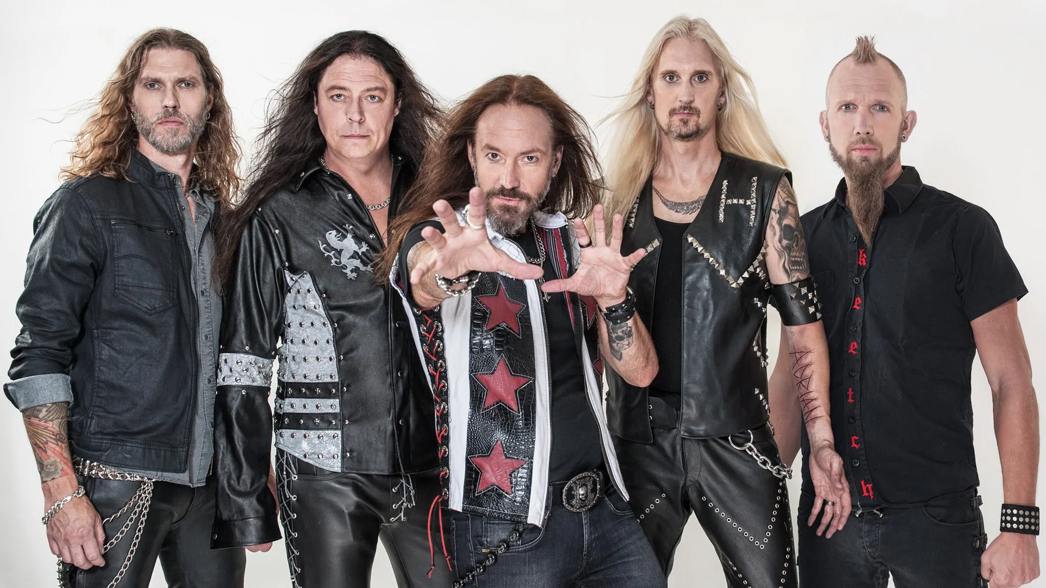 The five members of HammerFall in studded leather and denim posing against a white wall, the singer reaching toward the camera, ahead of their El Corazon show near Grappa