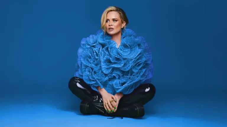 Comedian Joanne McNally sitting cross-legged in a large blue ruffled top and black patent trousers against a blue backdrop, ahead of her show at The Crocodile near Grappa