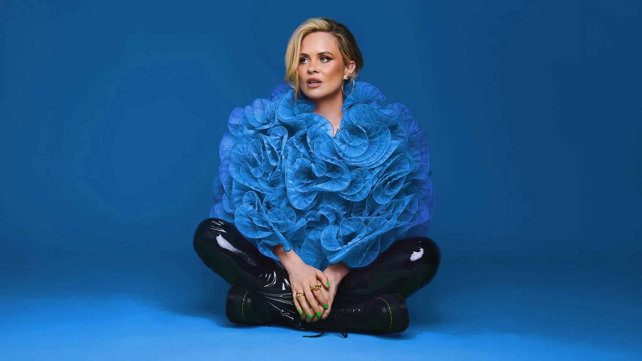 Comedian Joanne McNally sitting cross-legged in a large blue ruffled top and black patent trousers against a blue backdrop, ahead of her show at The Crocodile near Grappa
