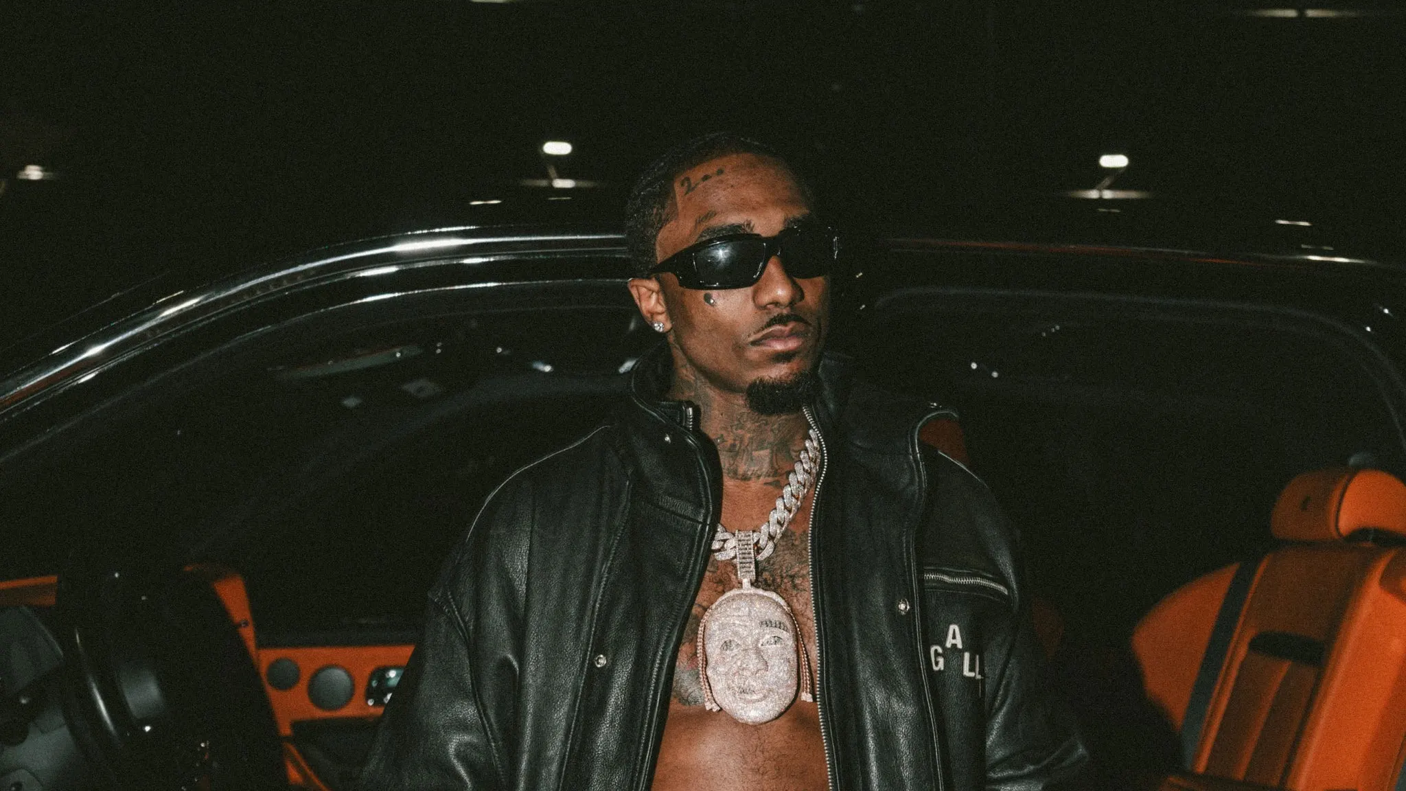 Rapper Loe Shimmy in dark sunglasses, a black leather jacket and a diamond pendant, standing by a car with orange leather seats, ahead of his show at The Crocodile near Grappa