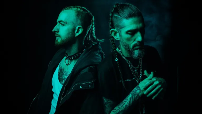 The two members of MISSIO, both with braided hair and tattoos, standing back to back in teal light against a dark background, ahead of their Funhouse show near Grappa