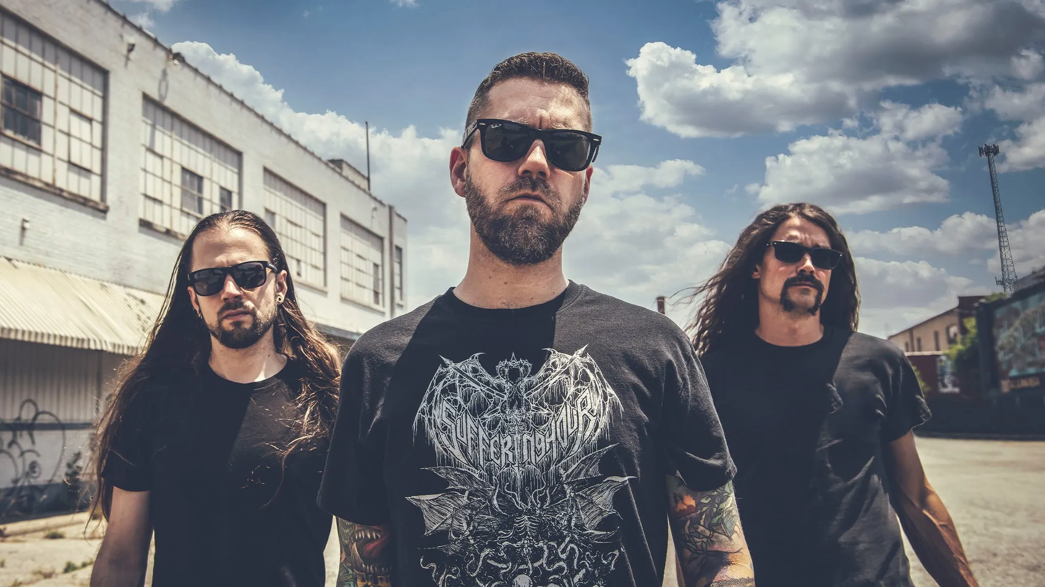 The three members of Revocation in sunglasses and black T-shirts standing on an empty street beside an old brick warehouse, ahead of their El Corazon show near Grappa