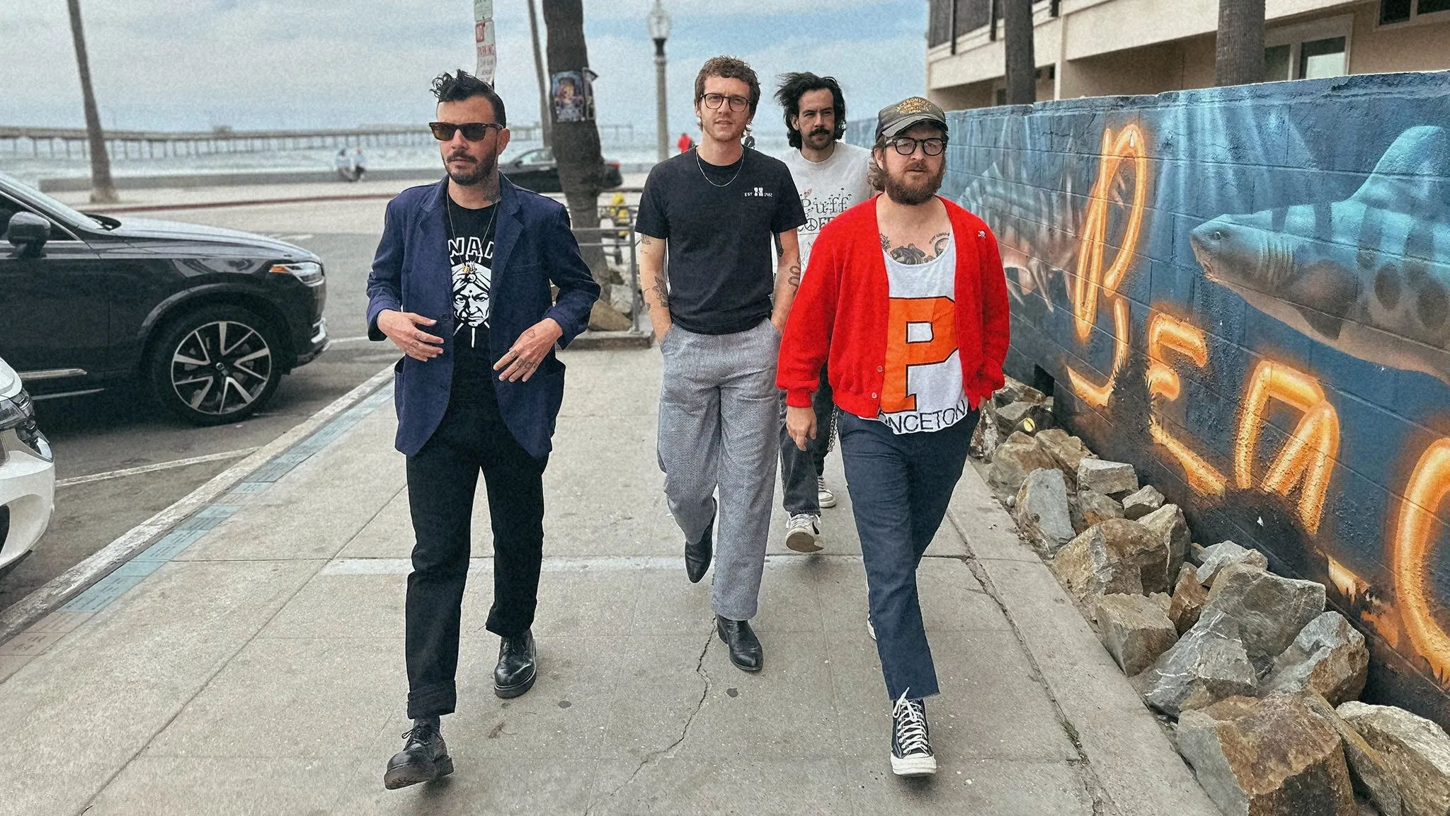 The four members of The Frights walking along a beachfront sidewalk past a mural of a shark on a cloudy day, ahead of their show at The Crocodile near Grappa
