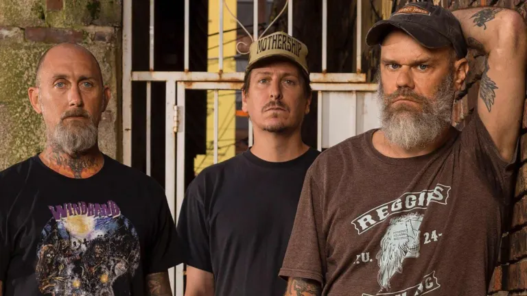 Three bearded band members in caps and T-shirts standing by a barred gate and a brick wall, promoting Weedeater and ZEKE at El Corazon near Grappa